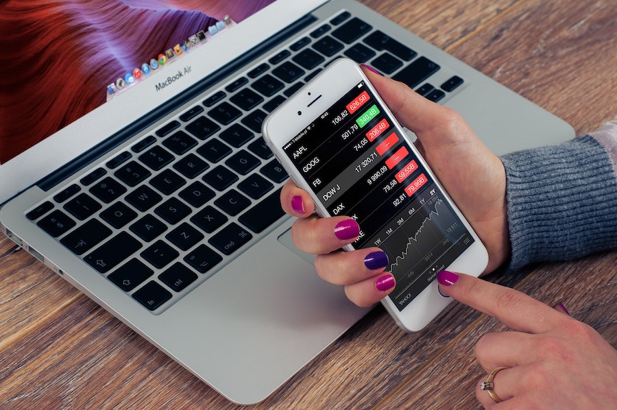 Woman with painted nails leaning on a MacBook whilst looking at Stock on an iPhone